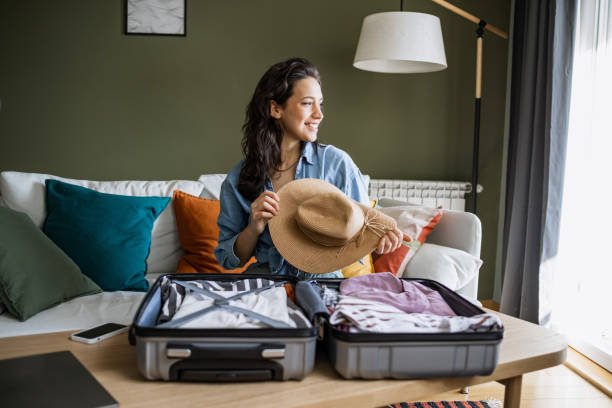 Young woman sitting on the couch and packing her travel suitcase. She is holding a straw hat in her hands