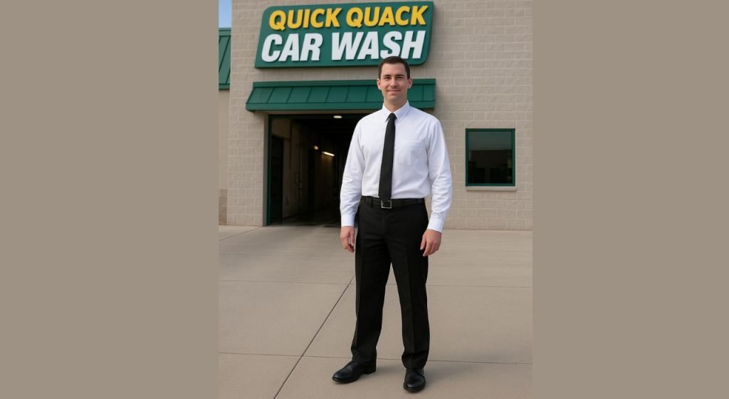 Male Quick Quack Car Wash employee wearing the official dress code — white button-down shirt, black tie, and black trousers — standing outside the Quick Quack Car Wash building.